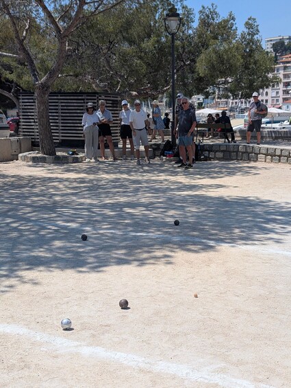 A group of people playing bocce in a park with trees and a lamp post. 4