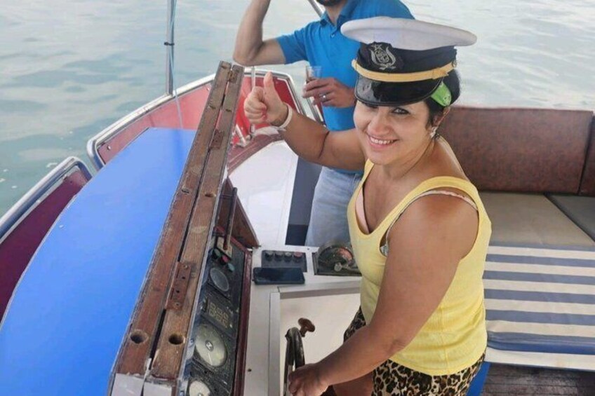 A person in a yellow top and patterned shorts at the helm of a boat with a blue and red hull. 1