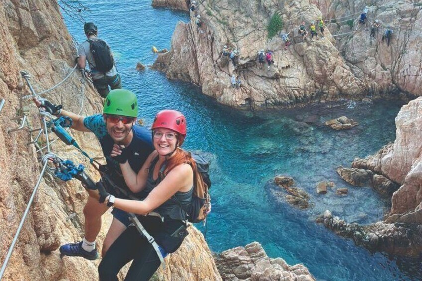 A couple rappelling down a rocky cliff with a clear blue sea in the background. 1