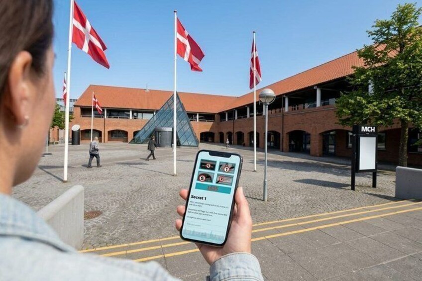 A person holding a smartphone with a travel app, standing in front of a building with Danish flags and a sign reading 'MCH'. 4