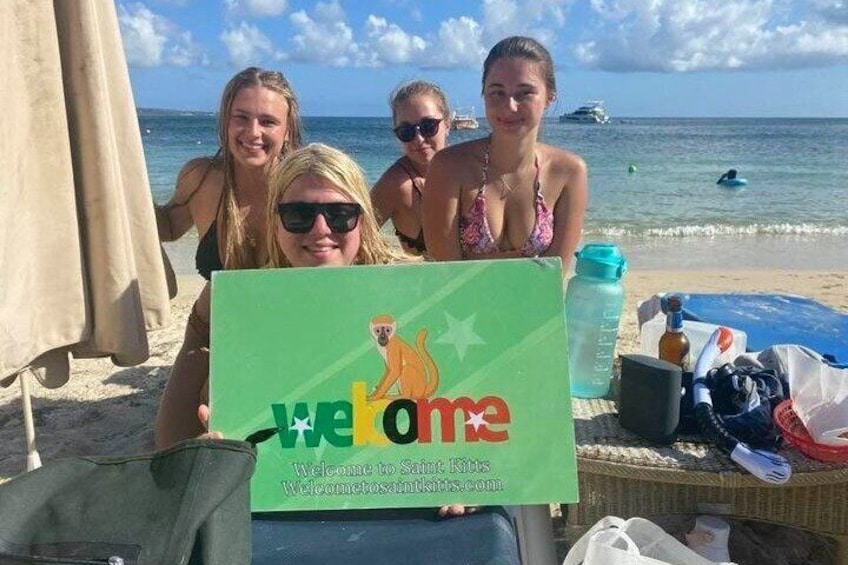A group of people holding a welcome sign on a beach with a clear sky and ocean in the background. 1