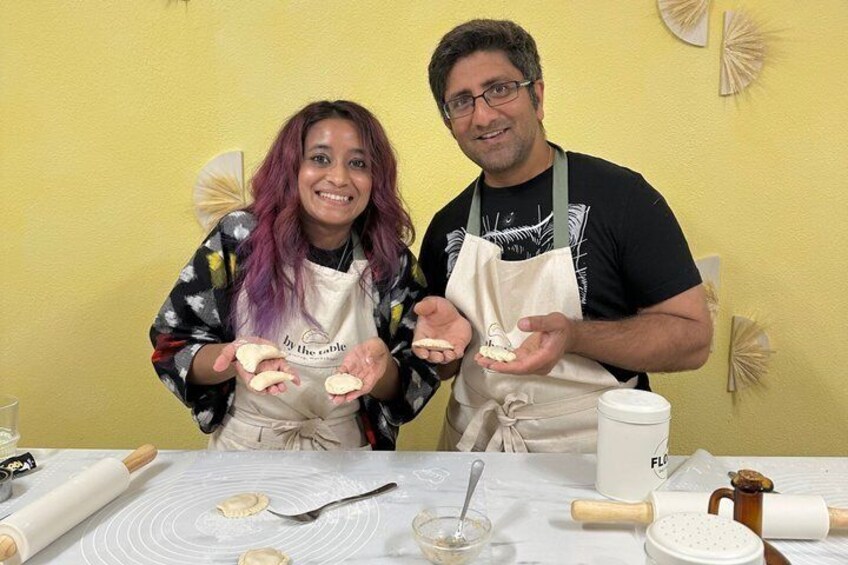 Two people in aprons holding dough, a rolling pin, a bowl, and a flour container on a table. 5