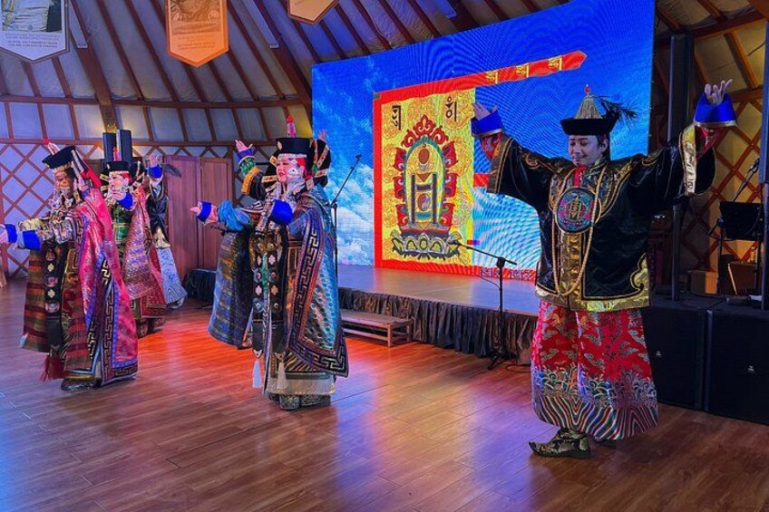 A stage with performers in traditional costumes, a large screen with a colorful backdrop, and a wooden floor. 1