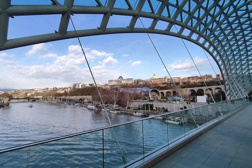 A modern bridge with a glass railing overlooking a river, with buildings and a dome in the background. 5