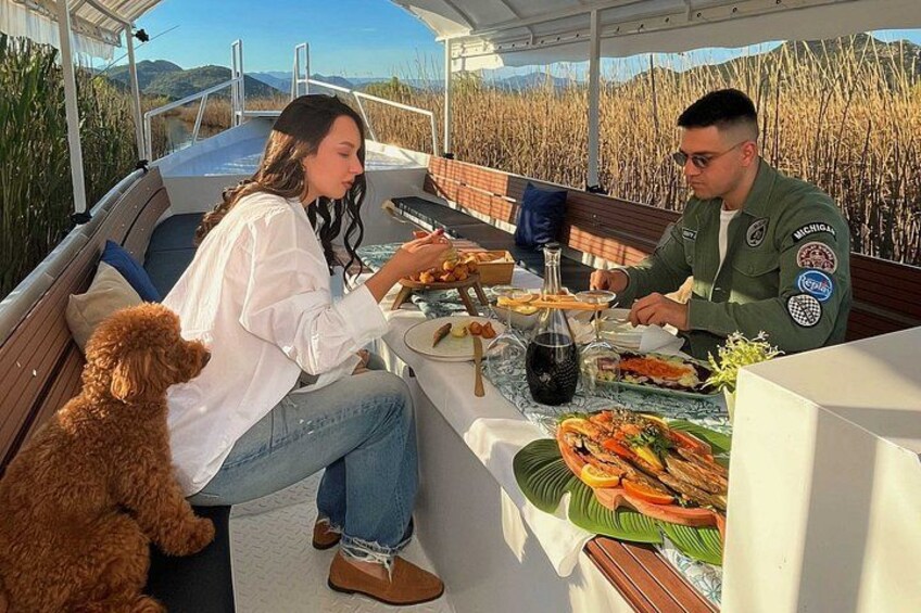 A boat with two people dining, a dog, and a scenic landscape. 1