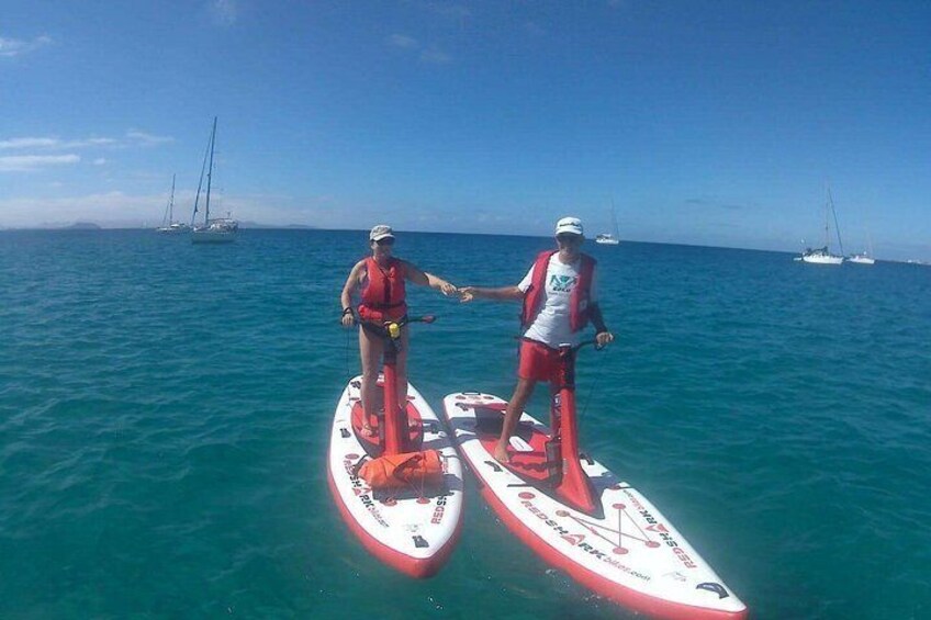 Two people paddleboarding on a calm sea with sailboats in the background. 2
