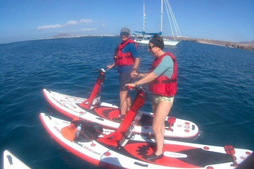 Two people on a red and white paddleboard in the water, with a sailboat in the background. 2