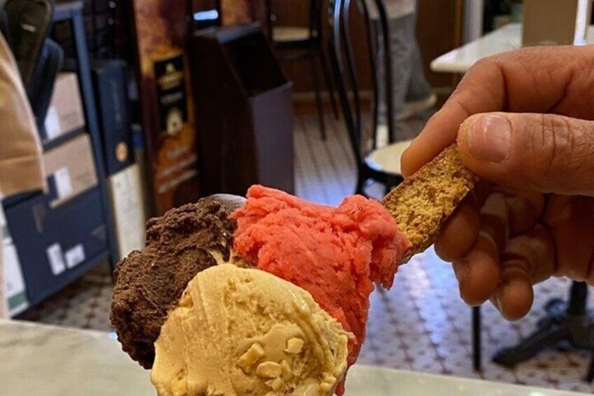 A hand holding a piece of chocolate with three scoops of ice cream in brown, red, and yellow colors. 3