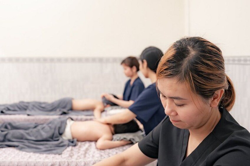 A massage therapy session with clients lying on treatment tables, a therapist attending to a client, and another person in the background. 3
