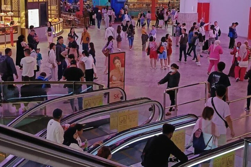 A busy indoor shopping mall with escalators, shoppers, and a prominent red advertisement. 2
