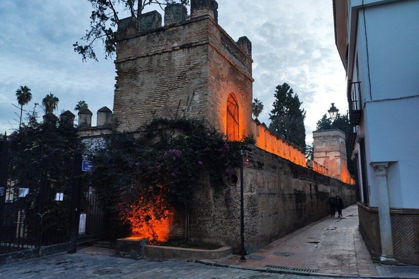 A historic stone wall with a lit-up window and a small garden area with flowers. 2