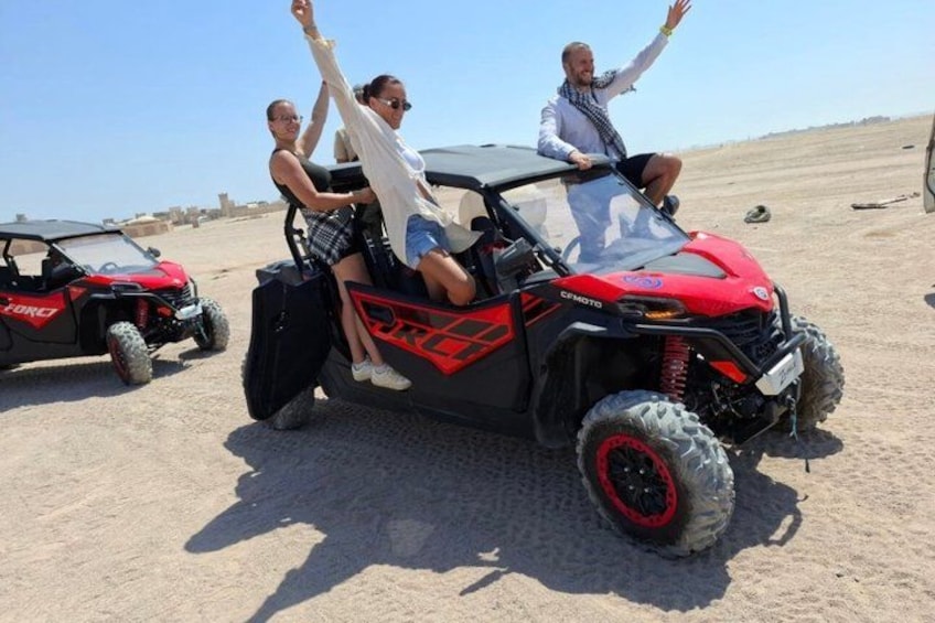 A red off-road vehicle with two passengers, a desert landscape, and another vehicle in the background. 4
