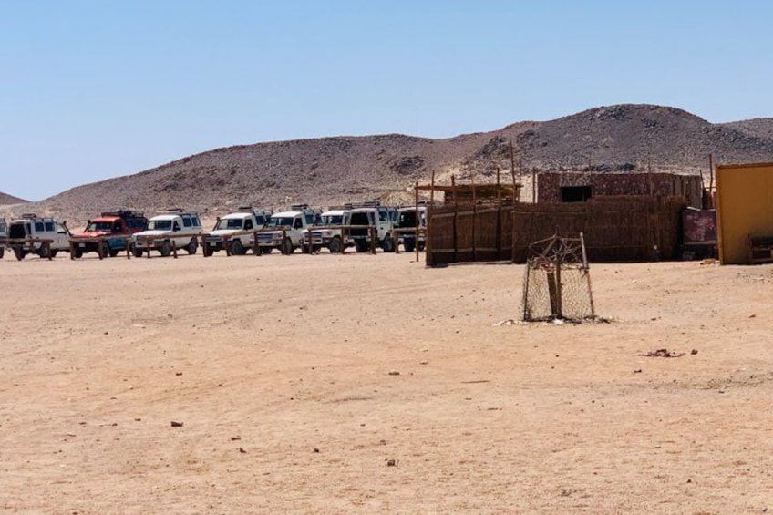A desert landscape with multiple vehicles parked in a row, a wooden structure, and a fenced area. 5
