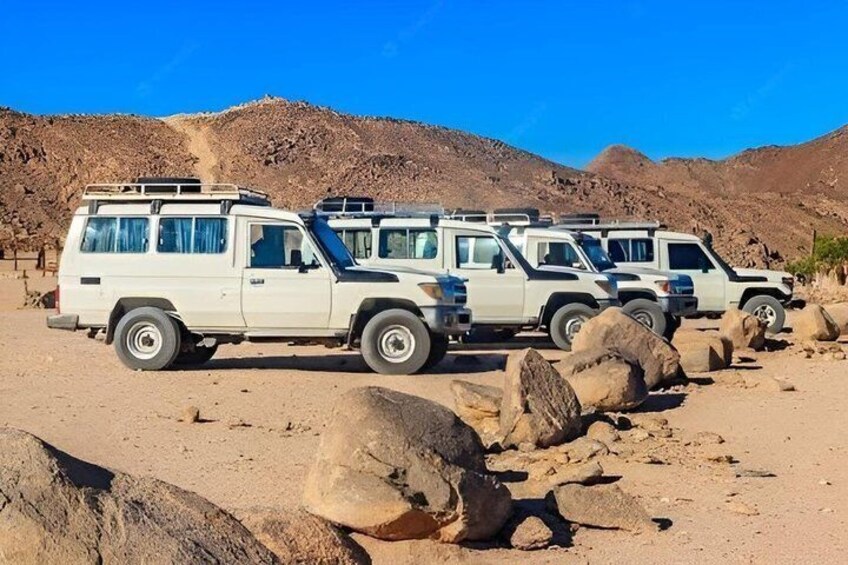 A row of white safari vehicles parked on a rocky terrain with large boulders in the foreground and a desert landscape in the background. 4