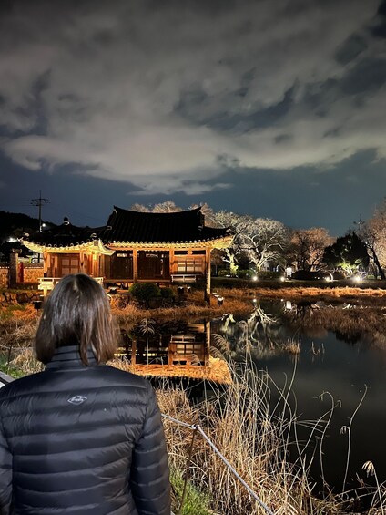 A person in a black jacket standing by a traditional building with a pond in the foreground. 4