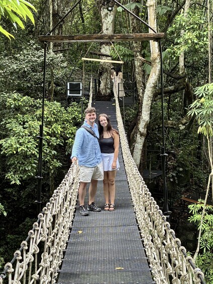 A couple standing on a rope bridge in a forested area with trees and a stone structure in the background. 4