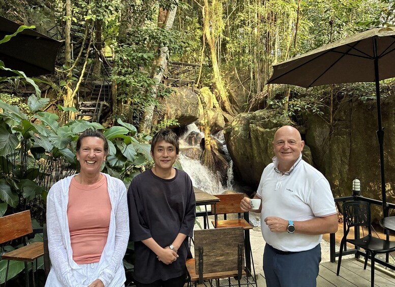 Three people standing in a garden with a waterfall and outdoor seating. 3