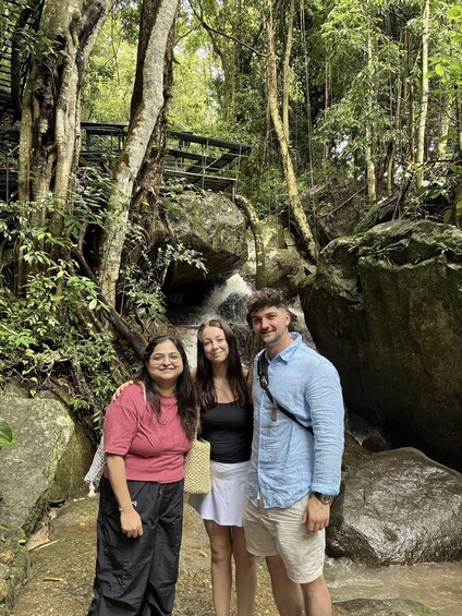 Three people standing in a forested area with a rock formation and a wooden bridge in the background. 3