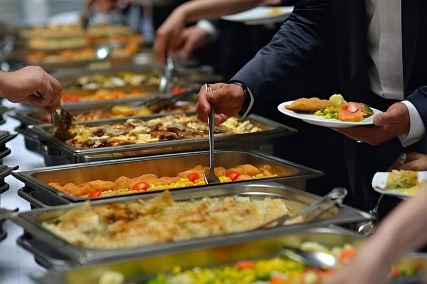 Un bufé con varios platos en bandejas, gente sirviéndose y una persona con traje sosteniendo un plato. 4