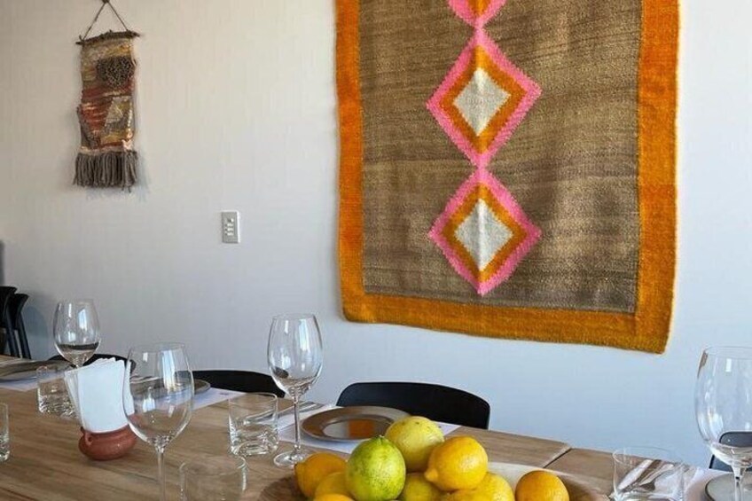 A dining table with a fruit bowl, wine glasses, and a decorative wall hanging. 4