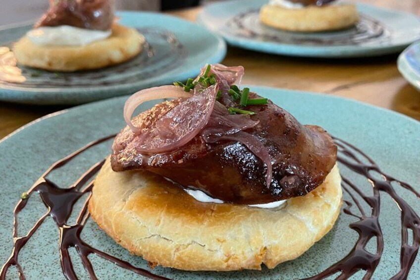 A dish with a glazed meat topping, garnished with onions and herbs, on a round bun with chocolate drizzle. 1