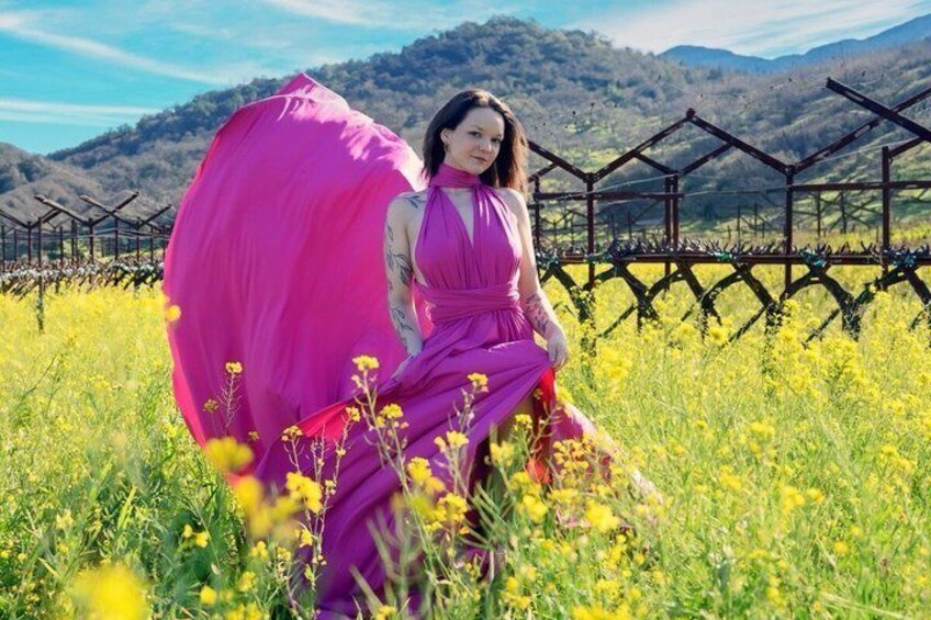 A person in a purple dress sitting in a field with yellow flowers and a vineyard in the background. 3