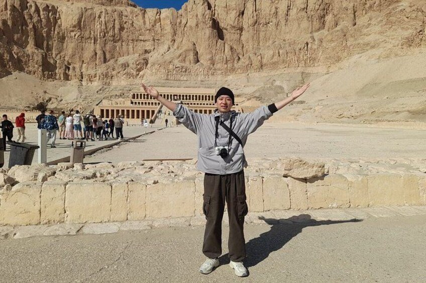 A person standing with arms outstretched in front of a large, ancient building with a desert backdrop. 2