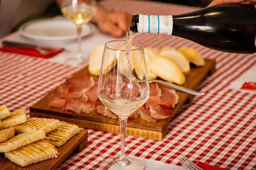 A bottle of wine being poured into a glass, with a checkered tablecloth, grilled bread, and sliced ham on a wooden board. 2