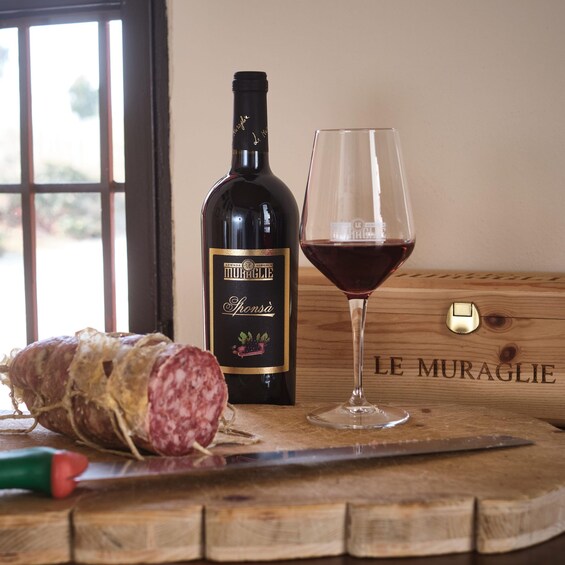 A bottle of wine, a glass of red wine, and a sliced salami on a wooden board with a knife. 4