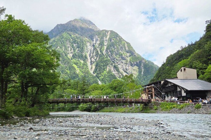 A wooden bridge over a river, a mountain in the background, and a building with a sloped roof. 1
