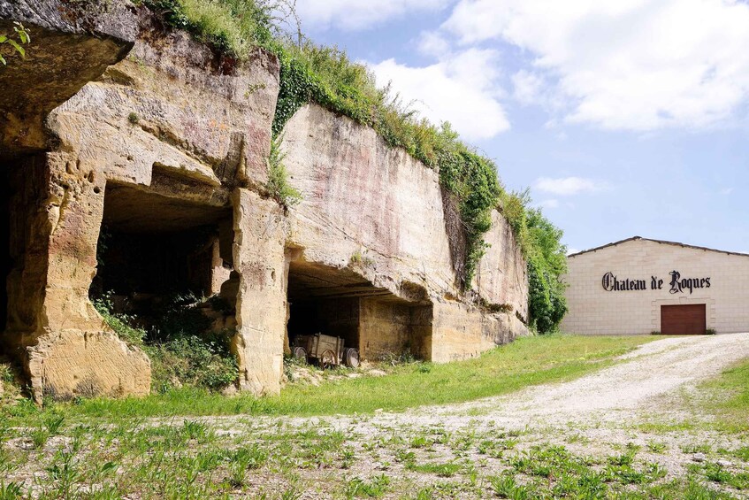 A building with 'Chateau de Roques' signage, a stone structure with openings, and a gravel path leading to it. 1