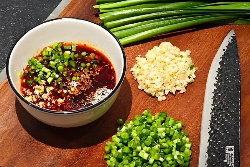 A bowl of sauce with chopped green onions, minced garlic, and a knife on a wooden surface. 5