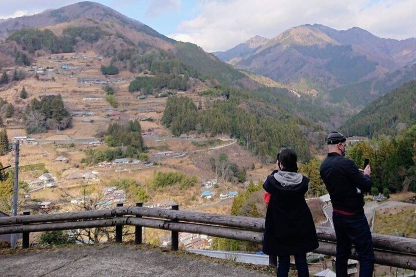 Two people standing on a wooden platform overlooking a valley with houses and mountains in the background. 2
