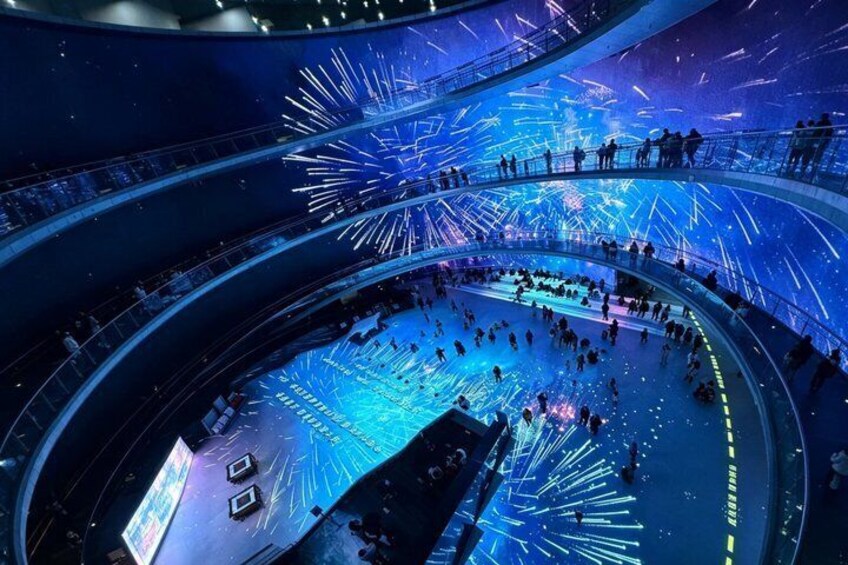A large indoor venue with a circular stage and audience seating, illuminated by blue and white lights. 4