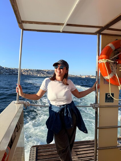 A person on a boat holding onto a pole, with a lifebuoy attached to the railing and a cityscape in the background. 2