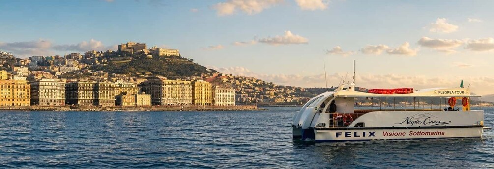 A boat named 'FELIX' with 'Naples Cruises' and 'Visione Sottomarina' on its side, sailing in the water with a cityscape and a hill with buildings in the background. 1