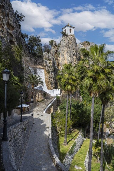 A stone pathway leading to a white bell tower atop a rocky cliff, surrounded by palm trees and greenery. 3