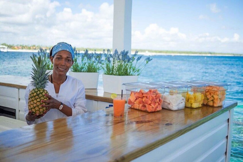 A person holding a pineapple at a buffet table with various fruits and a drink, overlooking a body of water. 4