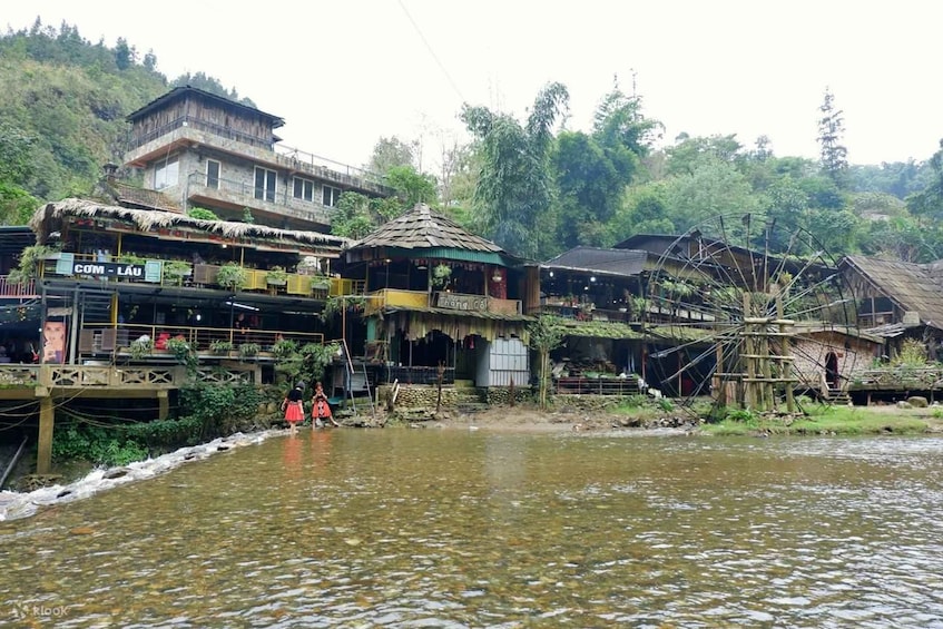 A riverside village with traditional buildings, a water wheel, and a sign for 'COM - LAU'. 4