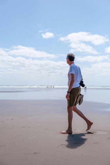 A man walking on a beach with a camera and flip-flops, a clear sky, and waves in the distance. 3