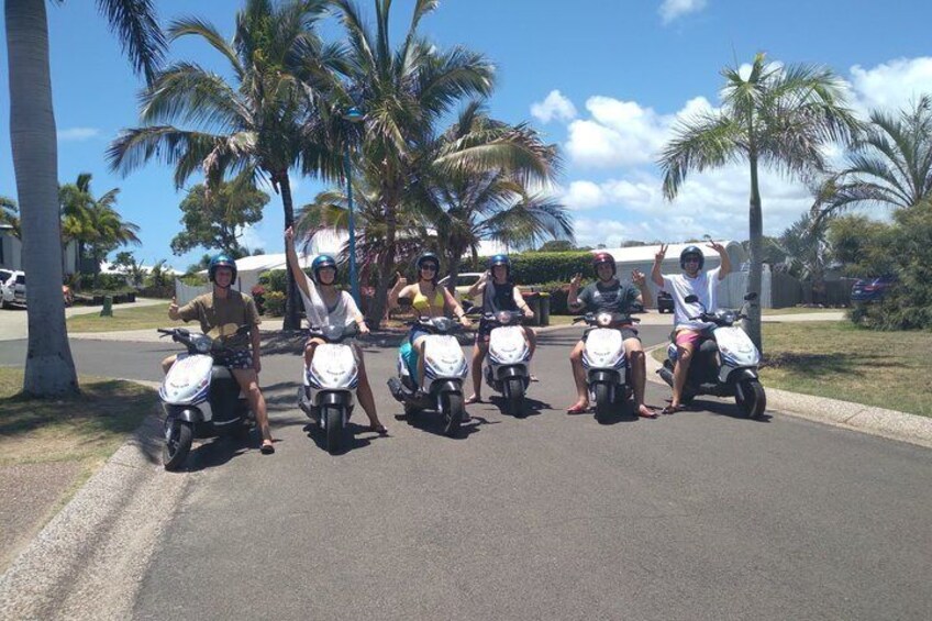Five people on scooters on a street lined with palm trees. 2
