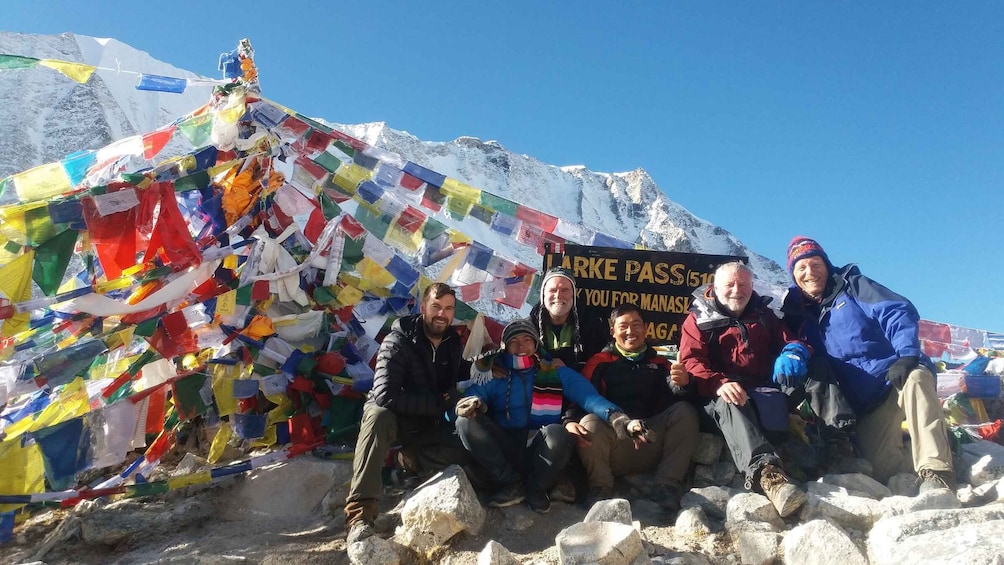 A group of people sitting on a snowy mountain with colorful prayer flags and a sign that reads "LARKE PASS". 1
