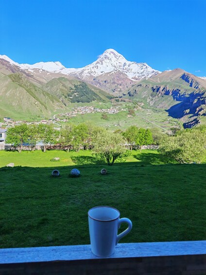 A mountain landscape with a village, a grassy field, and a white mug on a ledge. 4
