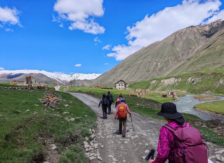 A group of hikers on a mountain trail with a river, a small building, and snow-capped mountains in the background. 5