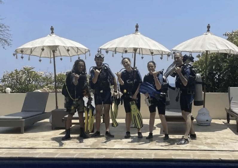 A group of people with scuba gear standing under umbrellas by a pool. 5