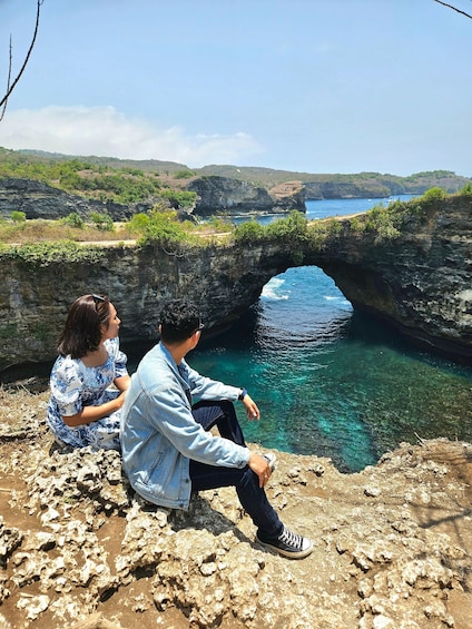 Dos personas sentadas sobre un afloramiento rocoso con vista a un mar azul y despejado y una formación rocosa natural. 2