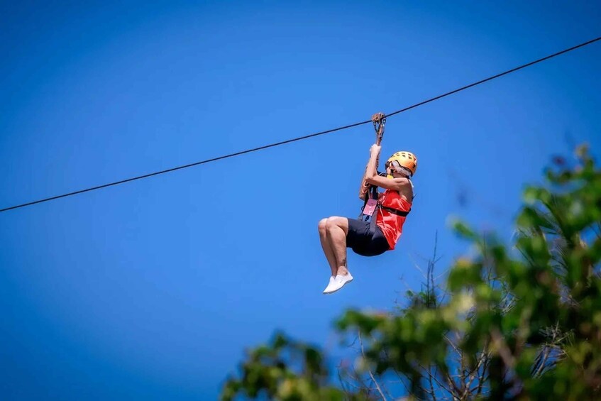 A person ziplining, wearing a helmet and harness, suspended on a cable against a clear blue sky. 1