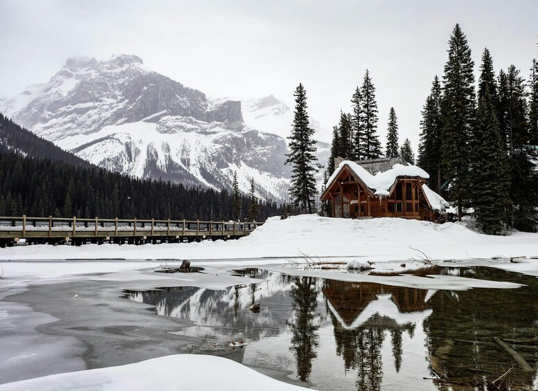 A wooden cabin surrounded by snow-covered mountains and a frozen lake with a reflection of the cabin and trees. 3