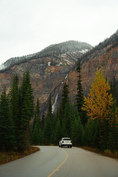 A winding road with a white SUV, surrounded by dense forests and a mountain with a waterfall. 4