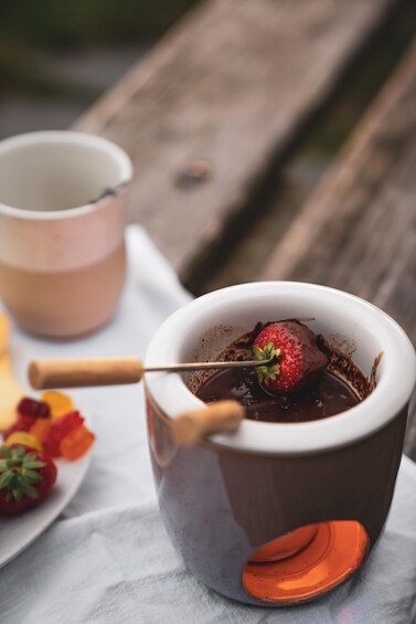 A dessert in a cup with a strawberry on top, a wooden spoon, and a plate with strawberries and a pink cup in the background. 3